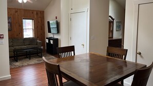 Un comedor con una mesa y sillas de madera, un televisor sobre un soporte, un sofá y una ventana con persianas.