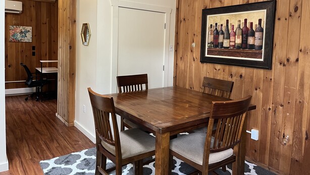 Un comedor con una mesa y sillas de madera, un cuadro de una botella de vino en la pared y una puerta que da a otra habitación.