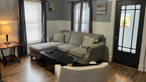 A living room with a sectional sofa, a coffee table, and a lamp. There is a door with a window, and a small table with a lamp. - Clinton House, Charming Cape Cod home decorated with flair in downtown Oneonta (Oneonta)