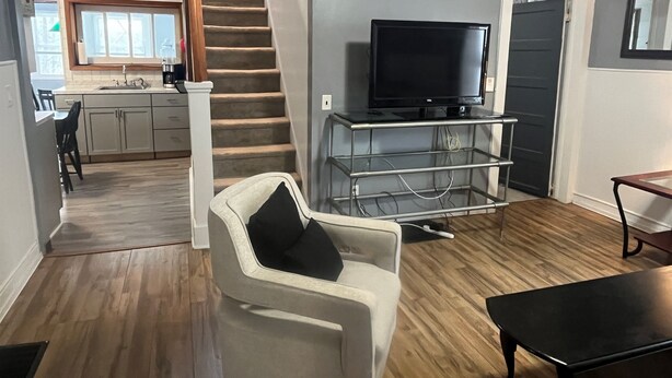 A living room with a TV on a stand, a white armchair, a wooden floor, and a staircase leading to a kitchen. - Clinton House, Charming Cape Cod home decorated with flair in downtown Oneonta (Oneonta)