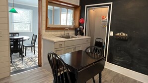 A modern kitchen with a dining area, featuring a black table and chairs, a window with blinds, and a hanging green light. - Clinton House, Charming Cape Cod home decorated with flair in downtown Oneonta (Oneonta)