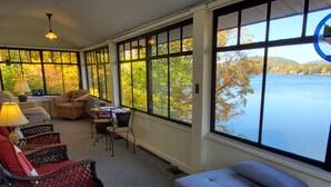 A sunlit room with a view of a lake, featuring a sofa, chairs, and a table. - Colone Cottage, Goodyear Lake, swimming, fishing, 3 kayaks, captivating view (Oneonta)