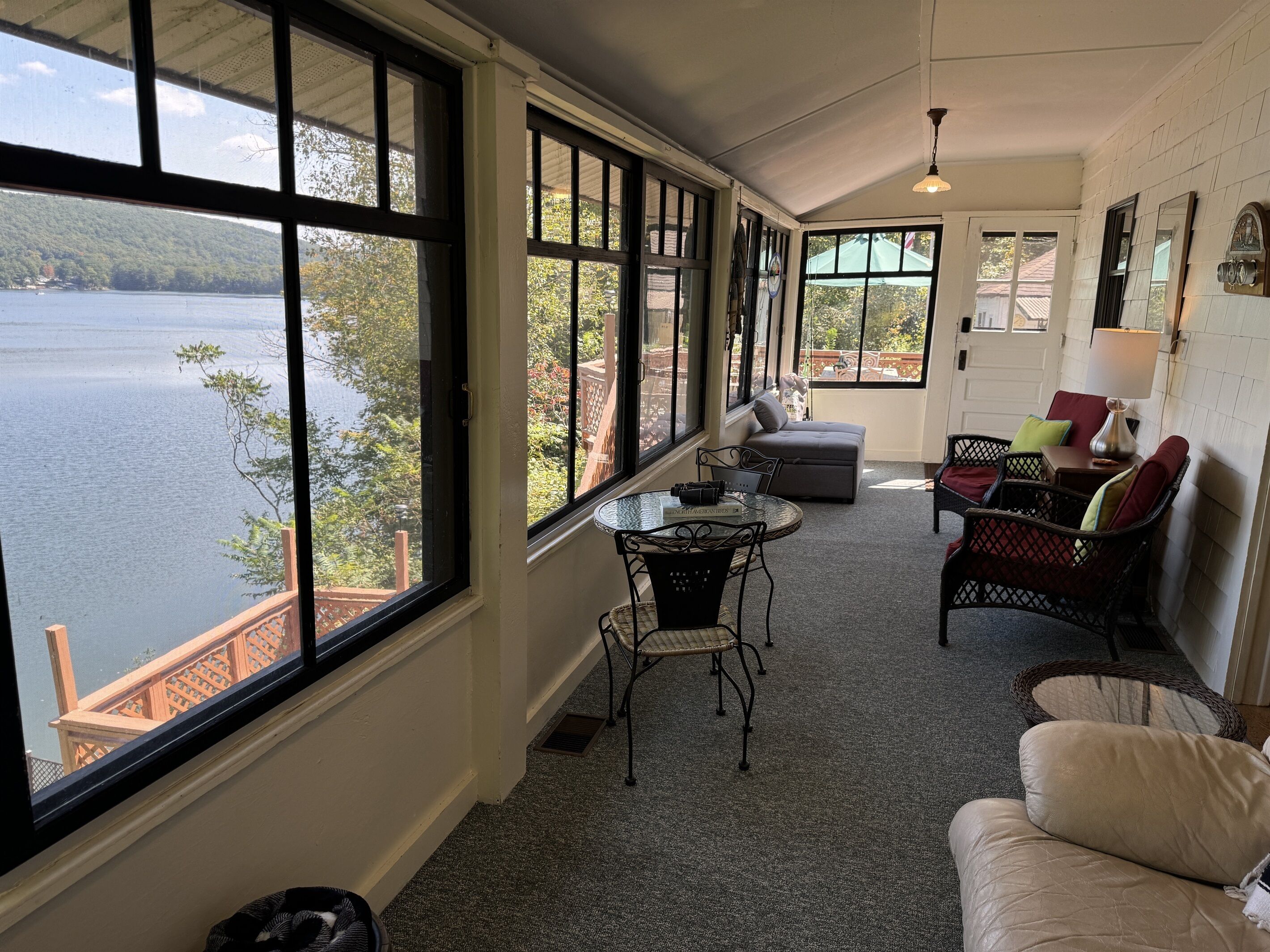 A sunlit room with large windows overlooking a lake, furnished with a table, chairs, and a sofa.