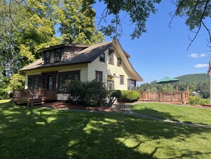 A two-story house with a porch, surrounded by a well-maintained lawn and trees. - Colone Cottage, Goodyear Lake, swimming, fishing, 3 kayaks, captivating view (Oneonta)