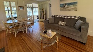 A living room with a gray sofa, a glass coffee table, and a dining area with a wooden table and white chairs. - Colone Cottage, Goodyear Lake, swimming, fishing, 3 kayaks, captivating view (Oneonta)