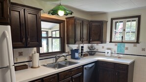 A kitchen with dark wooden cabinets, a white countertop, a built-in dishwasher, and a window with a view of trees. - Colone Cottage, Goodyear Lake, swimming, fishing, 3 kayaks, captivating view (Oneonta)