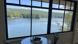 A room with a large window overlooking a lake, a round glass table with a book and a camera, and two wicker chairs. - Colone Cottage, Goodyear Lake, swimming, fishing, 3 kayaks, captivating view (Oneonta)