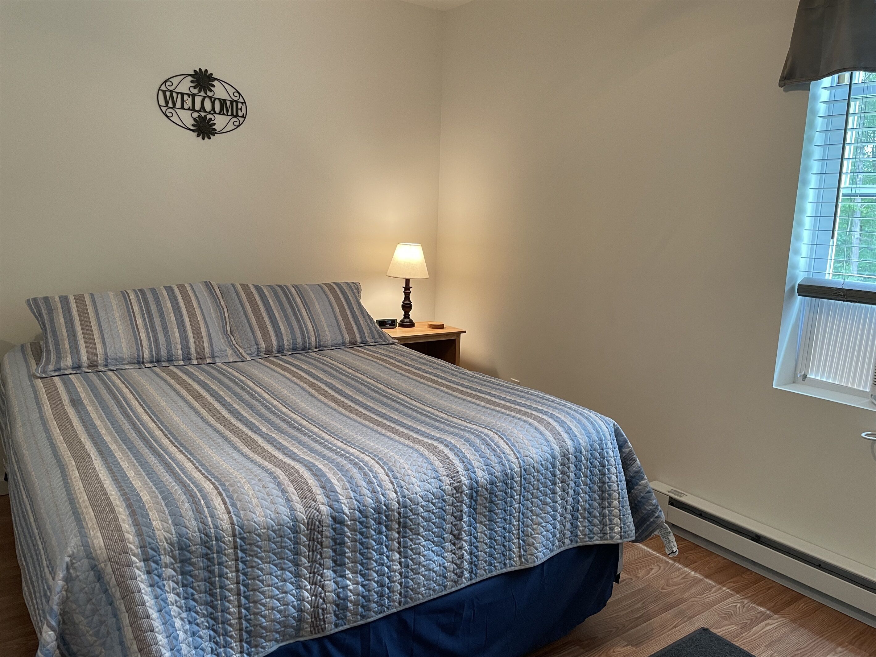 A bedroom with a bed, a nightstand, a lamp, a window, and a welcome sign on the wall.