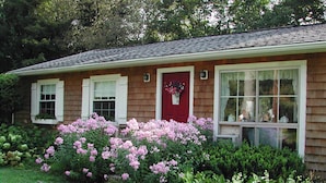 Ein Haus mit roter Tür, weißen Fenstern und einer Ziegel-Fassade. Vor der Tür befindet sich ein Garten mit rosa Blumen und grünen Sträuchern.