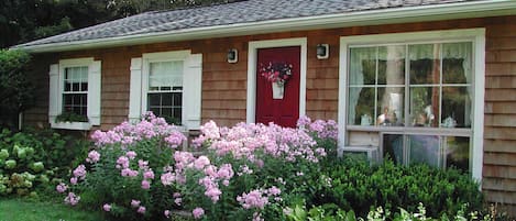 Una casa con puerta roja, ventanas blancas y fachada de ladrillo. En el frente, un jardín con flores rosadas y arbustos verdes.