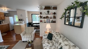 Ein Wohnzimmer mit floralem Sofa, gestreiftem Sessel und einem Couchtisch mit Dekoration. Im Hintergrund ist ein Essbereich mit Tisch und Stühlen zu sehen.