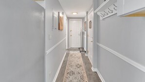 A hallway with light gray walls, a patterned rug, and a white door at the end. - #1 of 11 Units in Village of Cooperstown Apt Bldg (Cooperstown)