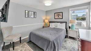 A bedroom with a bed, a chair, a desk, a TV, and a window with a view. - #1 of 11 Units in Village of Cooperstown Apt Bldg (Cooperstown)