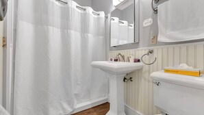 Un baño con cortina de ducha blanca, lavamanos pedestal, inodoro y espejo sobre el lavamanos.