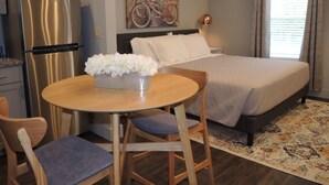 A compact hotel room with a bed, a dining table with chairs, a refrigerator, and a painting on the wall.