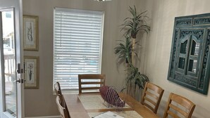 A dining area with a wooden table, chairs, a chandelier, a window with blinds, and framed artwork on the wall.