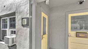 Rakennuksen ulkoseinä, jossa on keltainen ovi, ikkuna, jossa on turvakamera, ja ilmastointilaite.