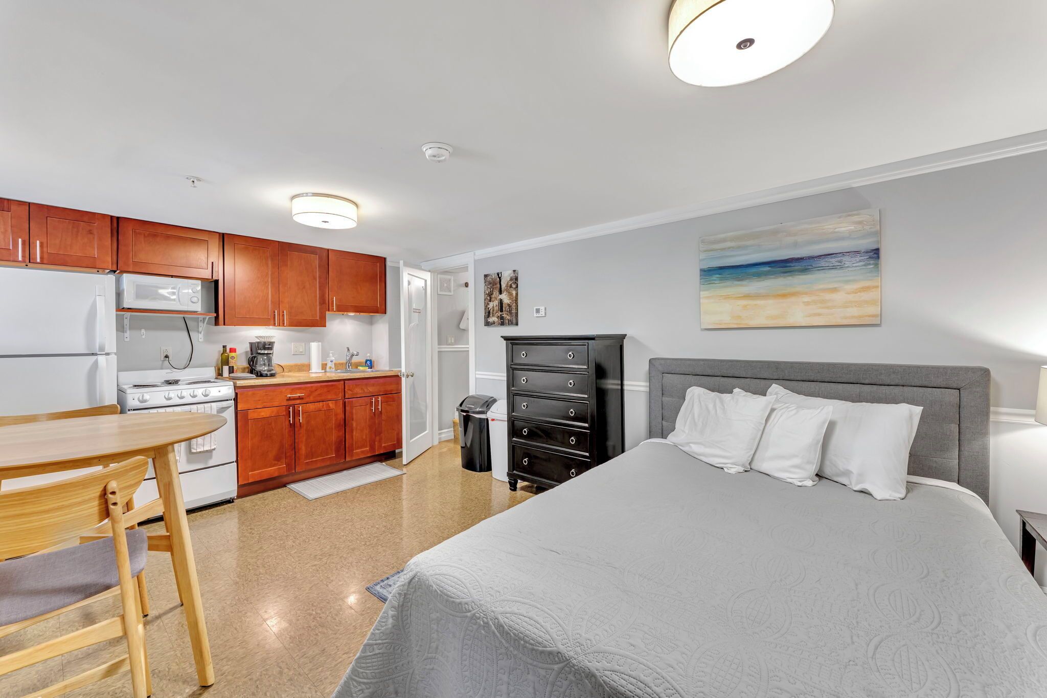 Una habitación de hotel compacta con kitchenette, una cama con cabecera gris y un cuadro en la pared.