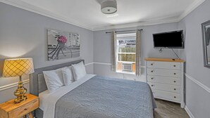 Un dormitorio con cama, mesita de noche con lámpara, cómoda, televisor montado en la pared y ventana con cortinas.