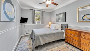 A bedroom with a bed, dresser, ceiling fan, artwork, and a window with curtains.