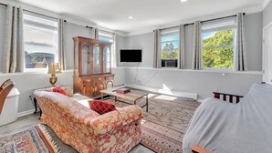 A spacious living room with a large window, a wooden cabinet, a flat-screen TV, and a patterned rug.
