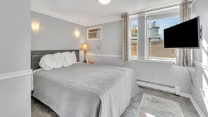 Un dormitorio con cama, televisor montado en la pared, ventana con cortinas, lámpara de noche y un cuadro pequeño en la pared.