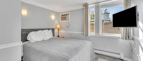 Um quarto com uma cama, televisão fixada na parede, janela com cortinas, abajur e uma pequena imagem na parede.