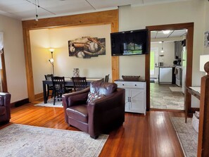 A living room with a leather sofa, a dining area with chairs and a table, a kitchen in the background, and a television mounted on the wall. - Diamond Dreams, Very close to Dreams Park, Recent update, fire pit, large yard (Cooperstown)
