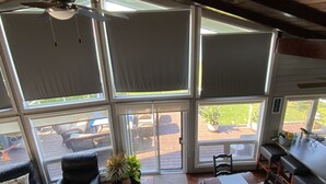 Ein geräumiges Zimmer mit einem großen Schiebeglasfenster, das auf eine Terrasse im Freien führt, einem Deckenventilator, einem Esstisch mit Stühlen und einem schwarzen Ledersofa.