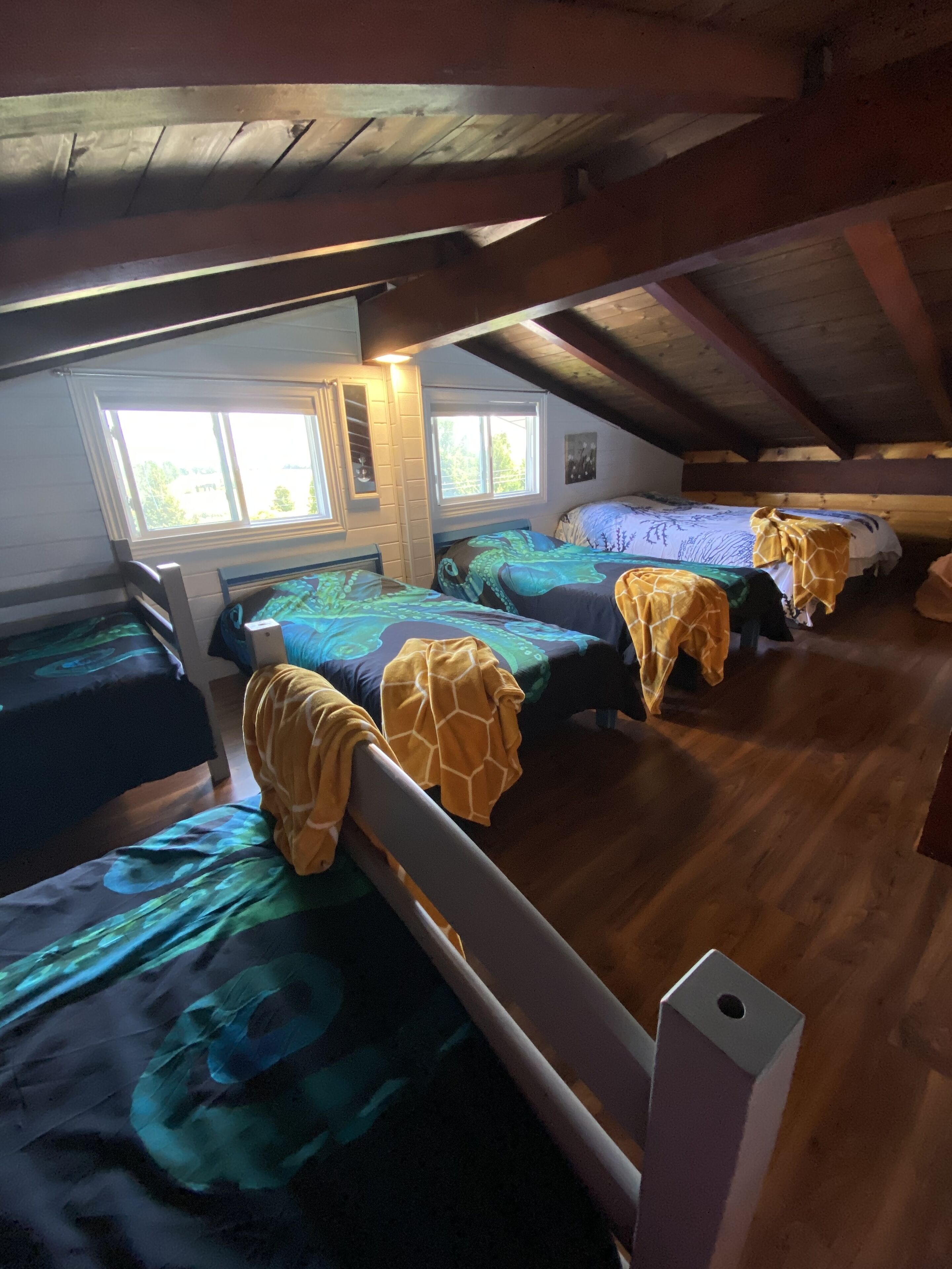 Habitación tipo dormitorio con cuatro camas dispuestas en fila, vigas de madera en el techo y una ventana que proporciona luz natural.