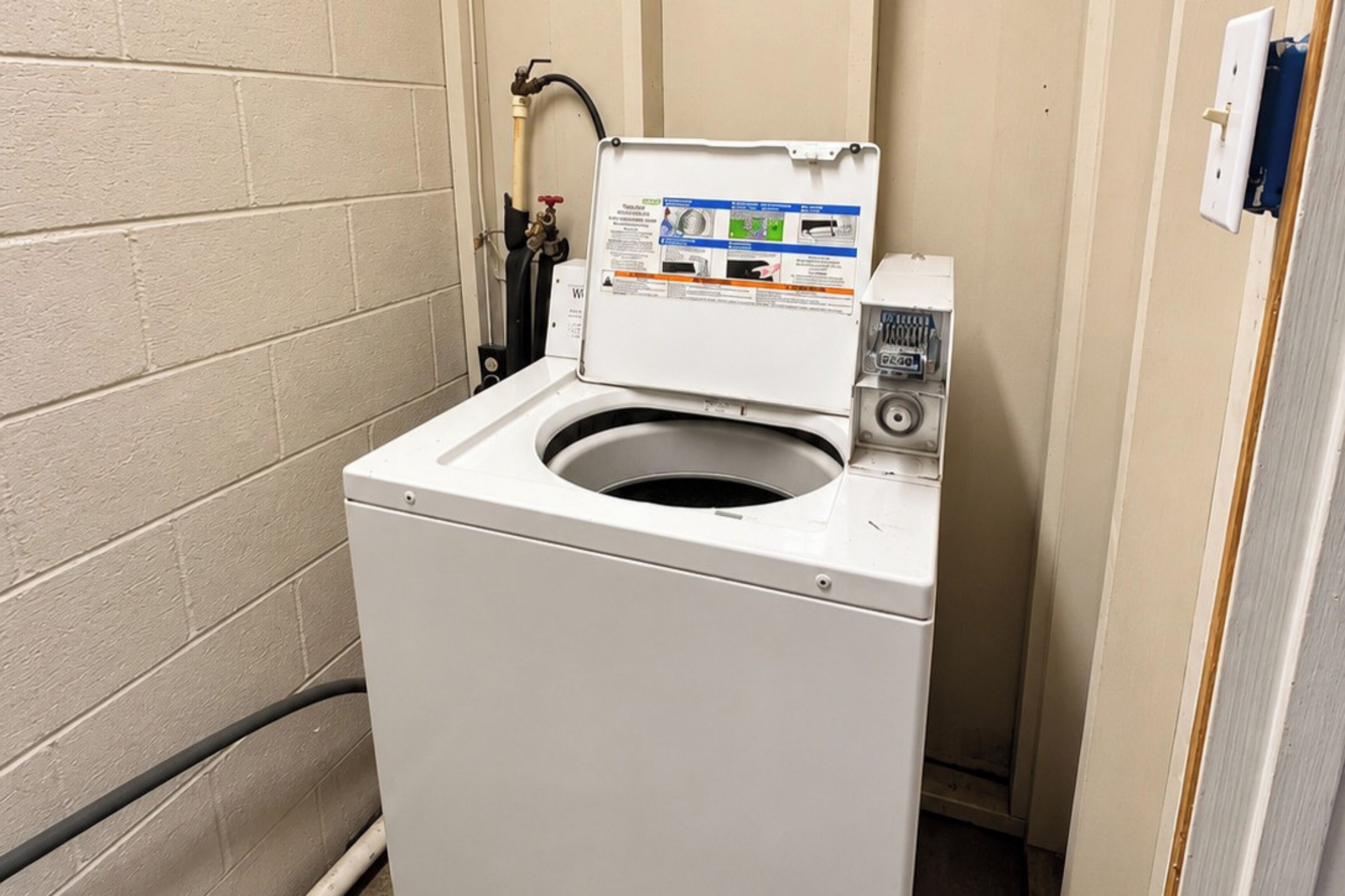Une machine à laver compacte, de couleur blanche, à chargement frontal, dotée d’un panneau de commande situé sur le dessus, et placée dans un coin d’une pièce aux murs en briques de couleur beige.