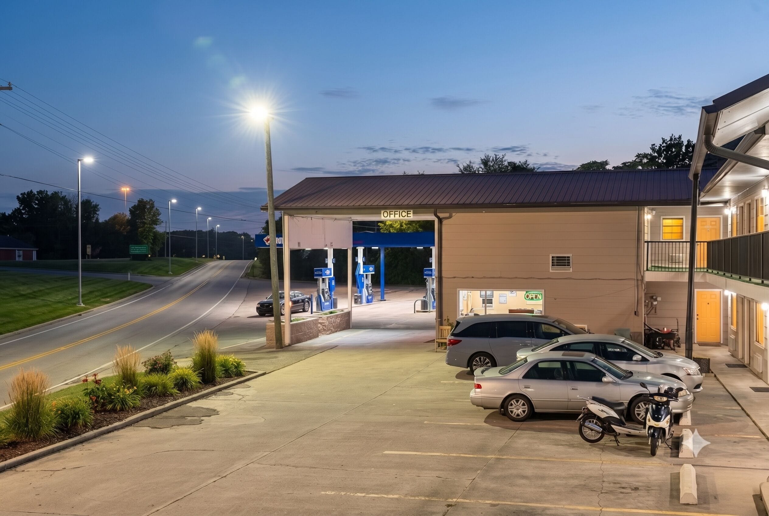 A gas station with a covered office area, multiple fuel pumps, and a parking lot with cars and a motorcycle.
