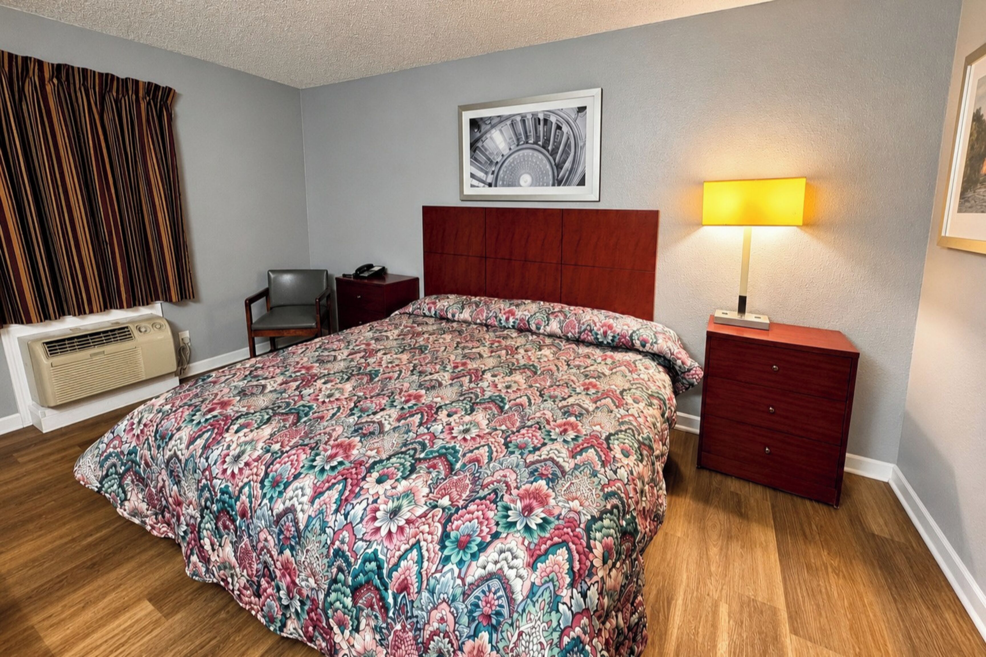 Une chambre d’hôtel avec un grand lit, une table de chevet avec une lampe, une chaise, un bureau avec un téléphone, un radiateur et un tableau encadré au mur.