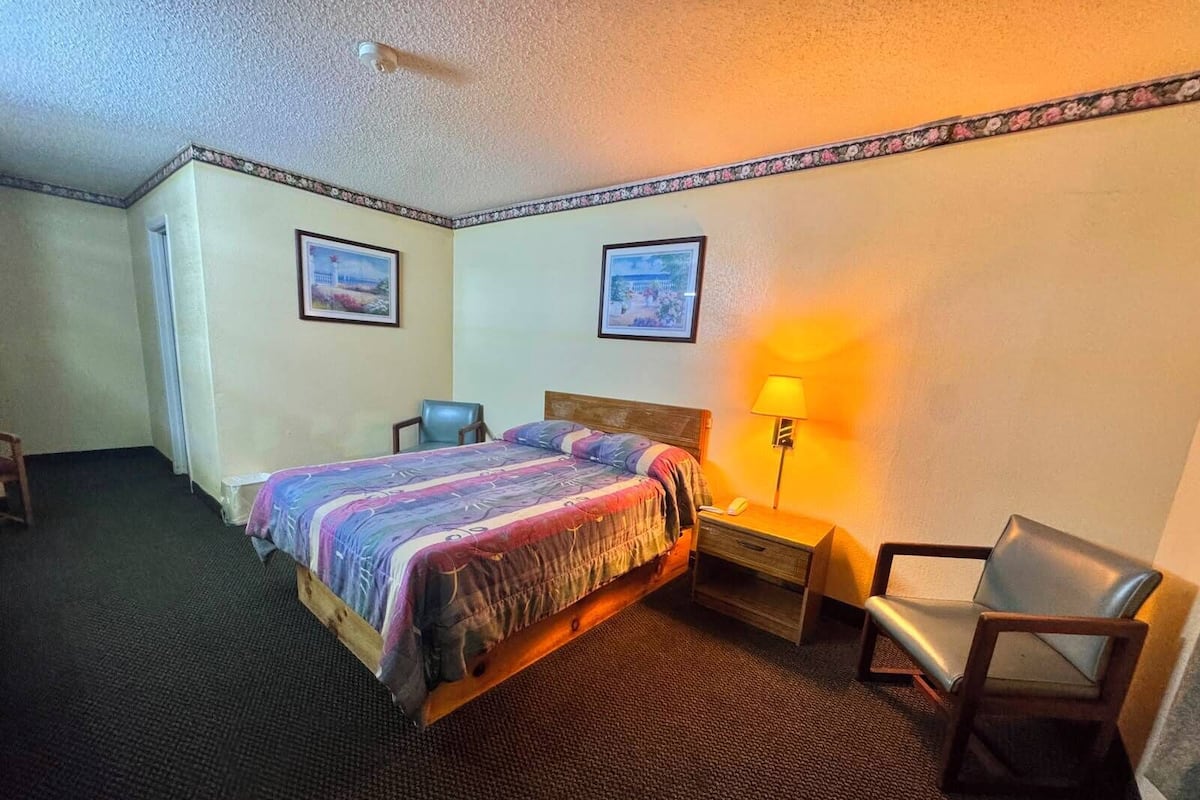 A hotel room with a bed, a chair, a desk with a lamp, and two framed pictures on the wall.