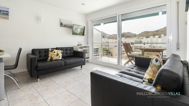 A modern living room with a black leather sofa, a dining table, and a large sliding glass door leading to a balcony with a city view. - Apartamento Watson by Mallorca North Villas (Port de Pollença)
