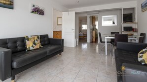 A modern living room with a black leather sofa, a dining area with a white table and chairs, and a kitchen area with a microwave and a TV. - Apartamento Watson by Mallorca North Villas (Port de Pollença)