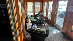 Hyggelig hytteinteriør med træloft, store vinduer, sofa, lænestol og lille bord.