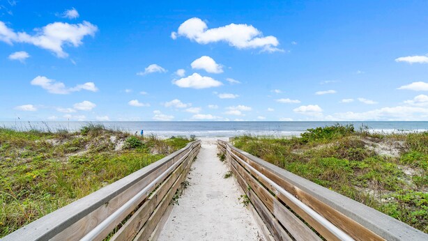 A sandy pathway leading to a beach with a clear sky and clouds.