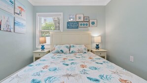 A bedroom with a bed, two bedside tables, a window with blinds, and wall decorations including framed pictures and a sign.