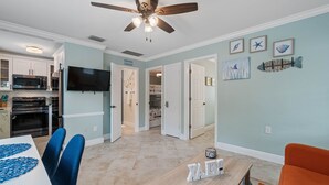 A spacious living area with a dining table, blue chairs, and a ceiling fan. There is a kitchen with modern appliances and a flat-screen TV.