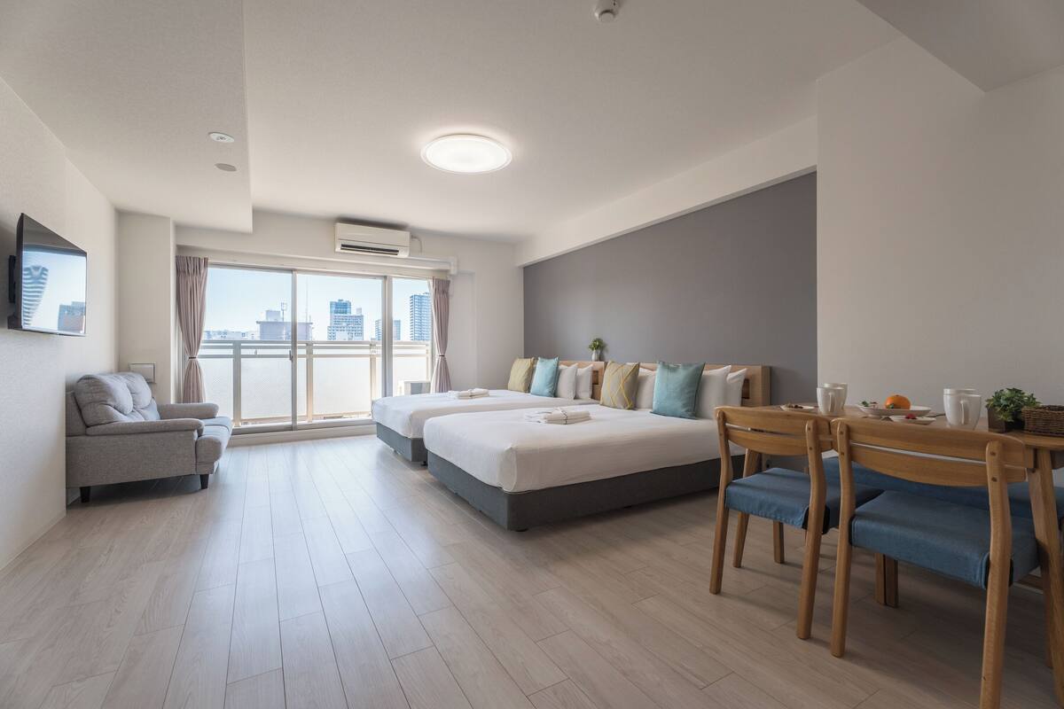 A modern hotel room with a large bed, a sofa, a dining table with chairs, and a city view through the window.