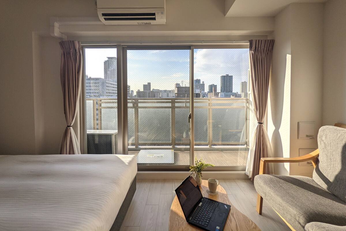 A hotel room with a bed, a desk with a laptop, a chair, and a large window with a city view.