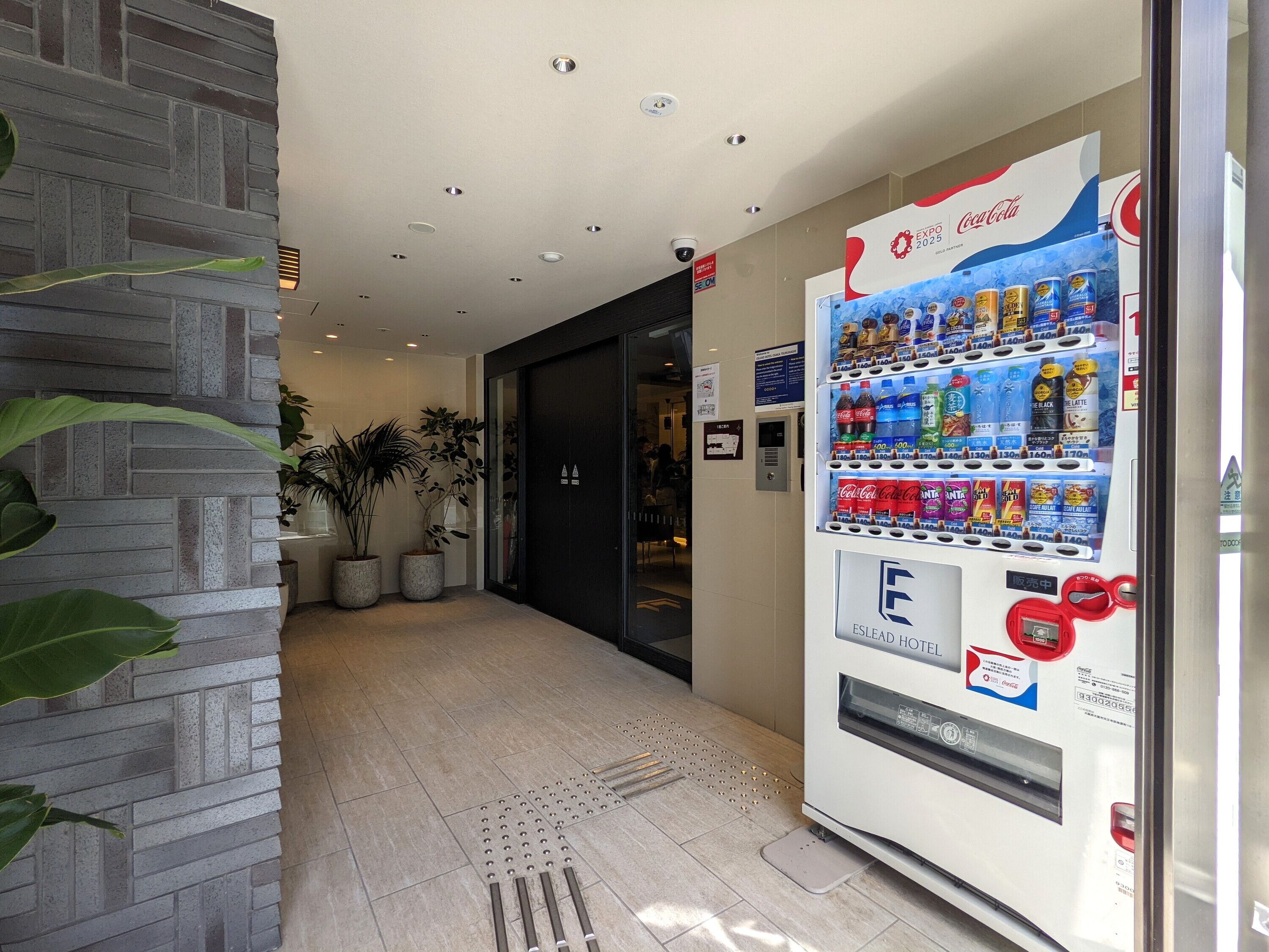 Máquina expendedora con diferentes bebidas y aperitivos, pared de ladrillo y plantas en macetas en un pasillo.