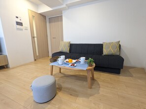 A modern living room with a wooden floor, a grey ottoman, a small coffee table with a plate of fruit, and a dark sofa with patterned cushions. - ESLEAD HOTEL OSAKA TSURUHASHI (Osaka)