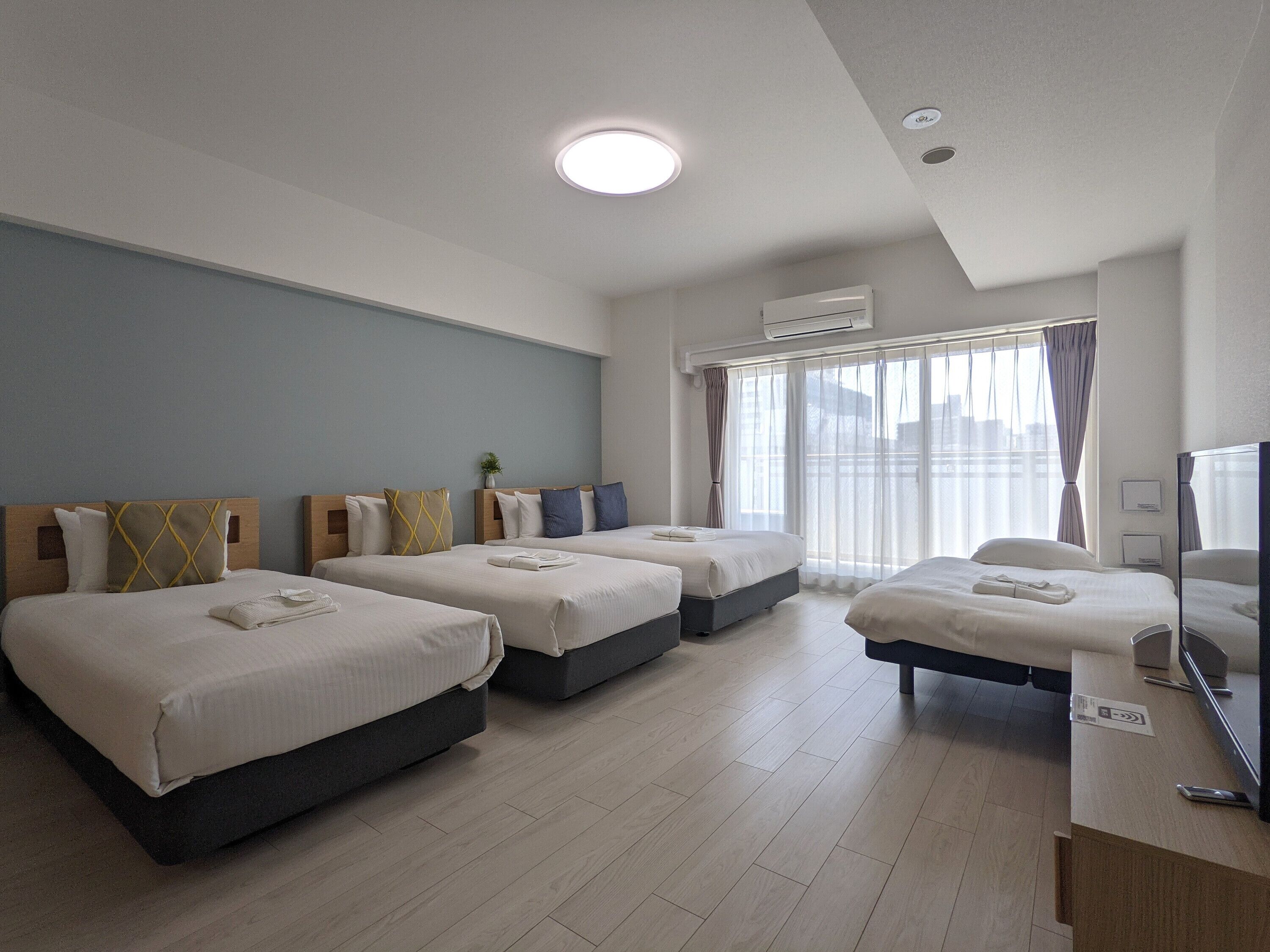 Habitación de hotel con tres camas, televisión de pantalla plana y un ventanal grande con cortinas.