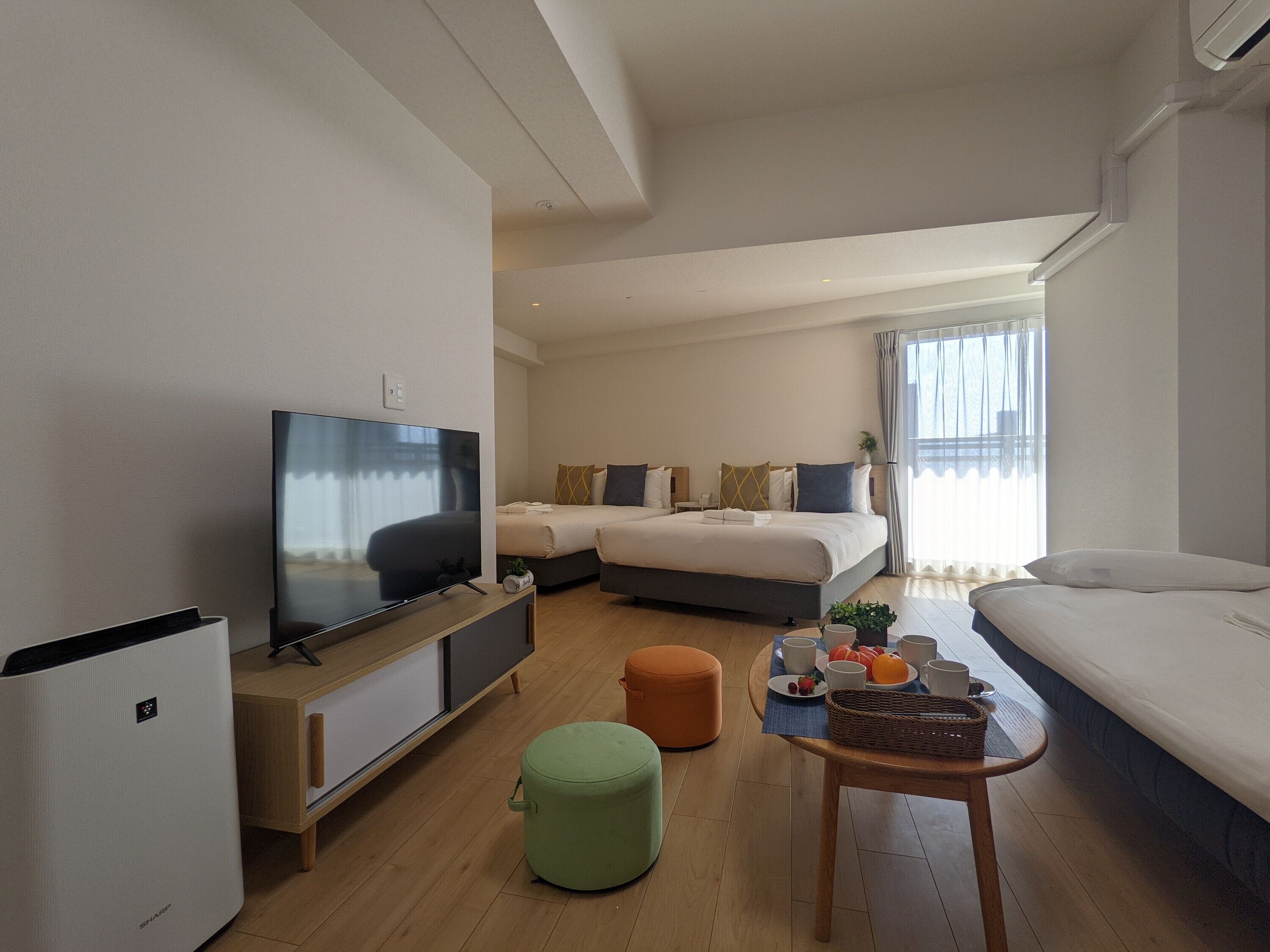 A modern hotel room with a flat-screen TV, a sofa, a bed with multiple pillows, a small table with a basket, and a window with curtains.