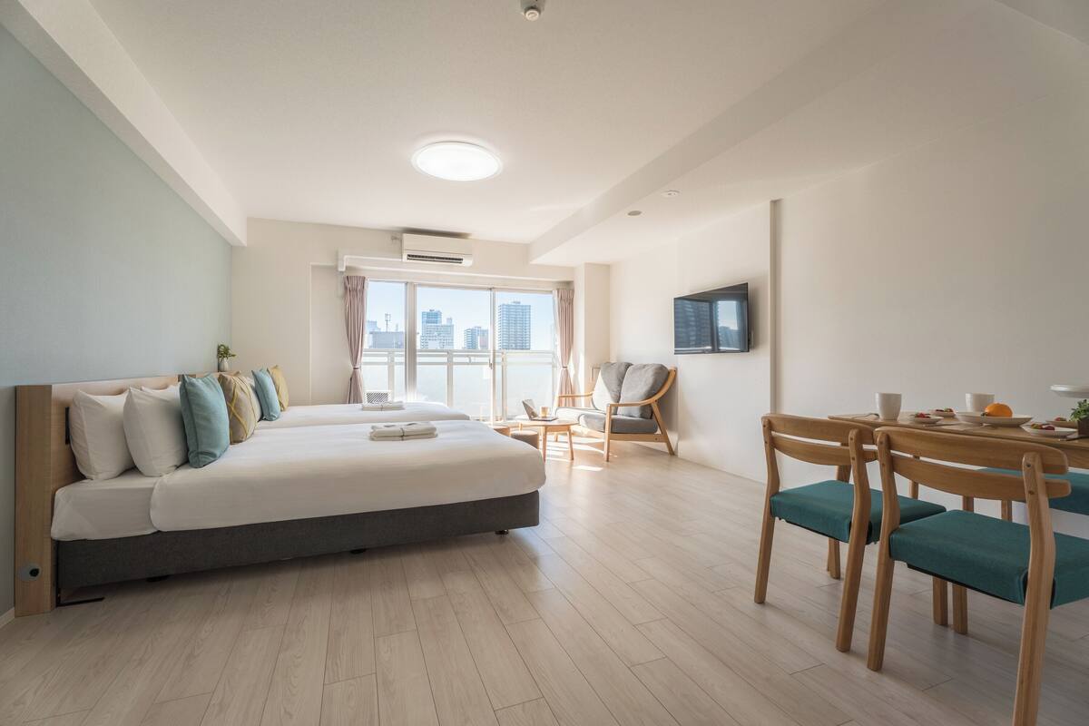 A modern hotel room with a large bed, a dining table with chairs, and a city view through the window.