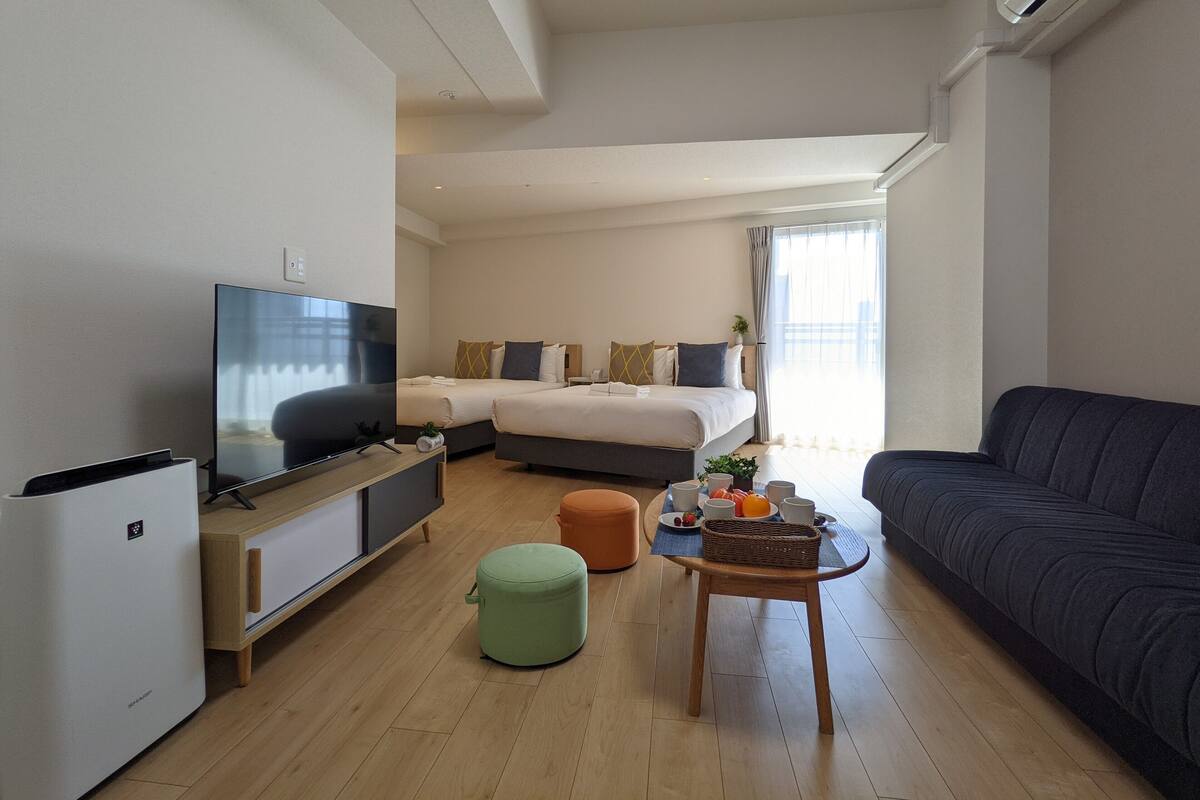 A modern hotel room with a flat-screen TV, a sofa, a bed, a small table with a basket, and a green ottoman.