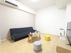 A modern living room with a dark grey sofa, a wooden coffee table with fruits and cups, a yellow ottoman, and a grey storage ottoman. - ESLEAD HOTEL OSAKA TSURUHASHI (Osaka)