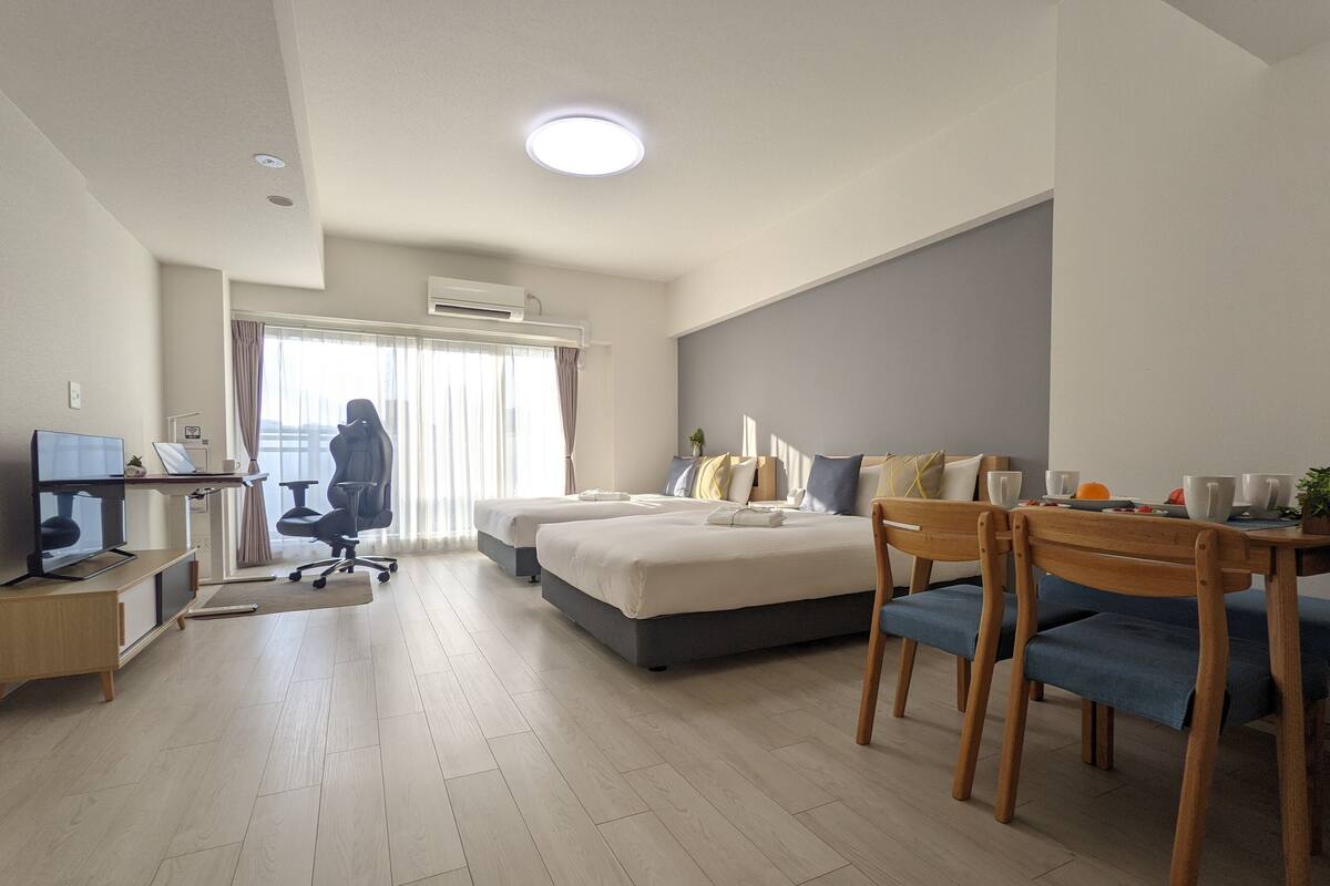 A modern hotel room with a desk, two beds, a dining area, and an air conditioning unit.